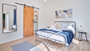 Une chambre à coucher moderne avec un cadre de lit en métal, une porte coulissante en bois, un grand miroir et des œuvres d’art abstraites sur les murs.