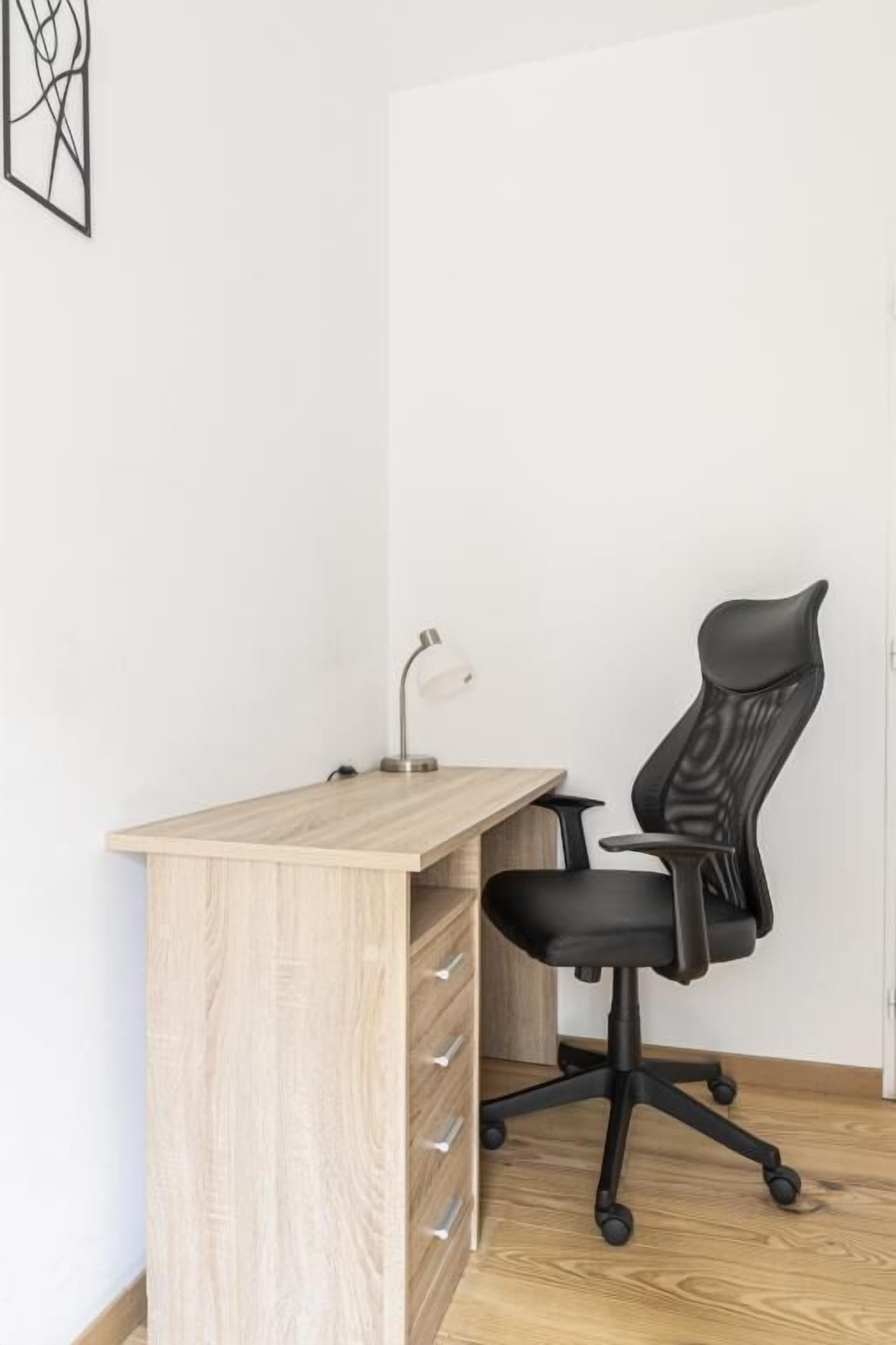 Een modern bureau met een houten afwerking, een zwarte bureaustoel en een bureaulamp.