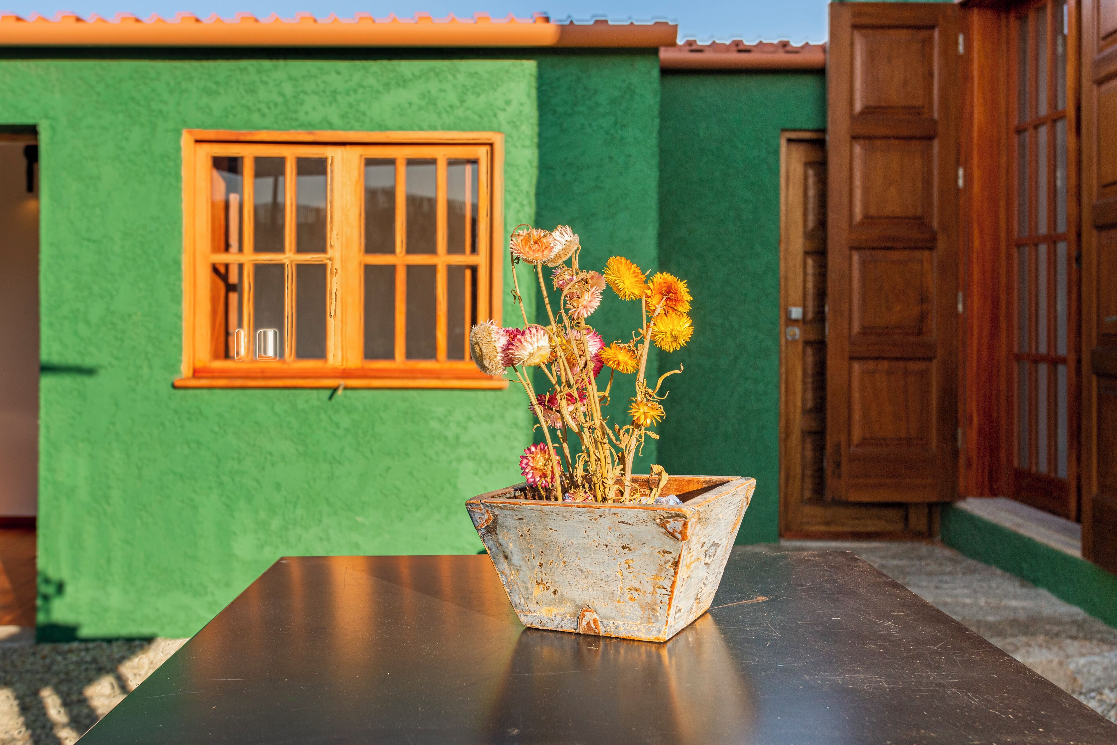 A rustic, weathered flower pot on a table in front of a green building with a wooden door and window.