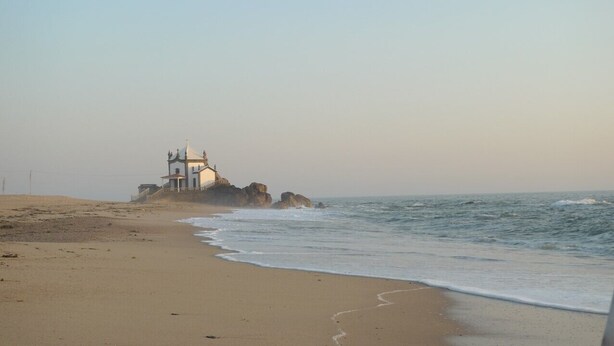 A beach with a small white building on a rock, a sandy shore, and the ocean.