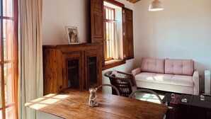 A cozy living room with a wooden ceiling, a white sofa, a wooden table with a glass vase, and a window with wooden shutters.