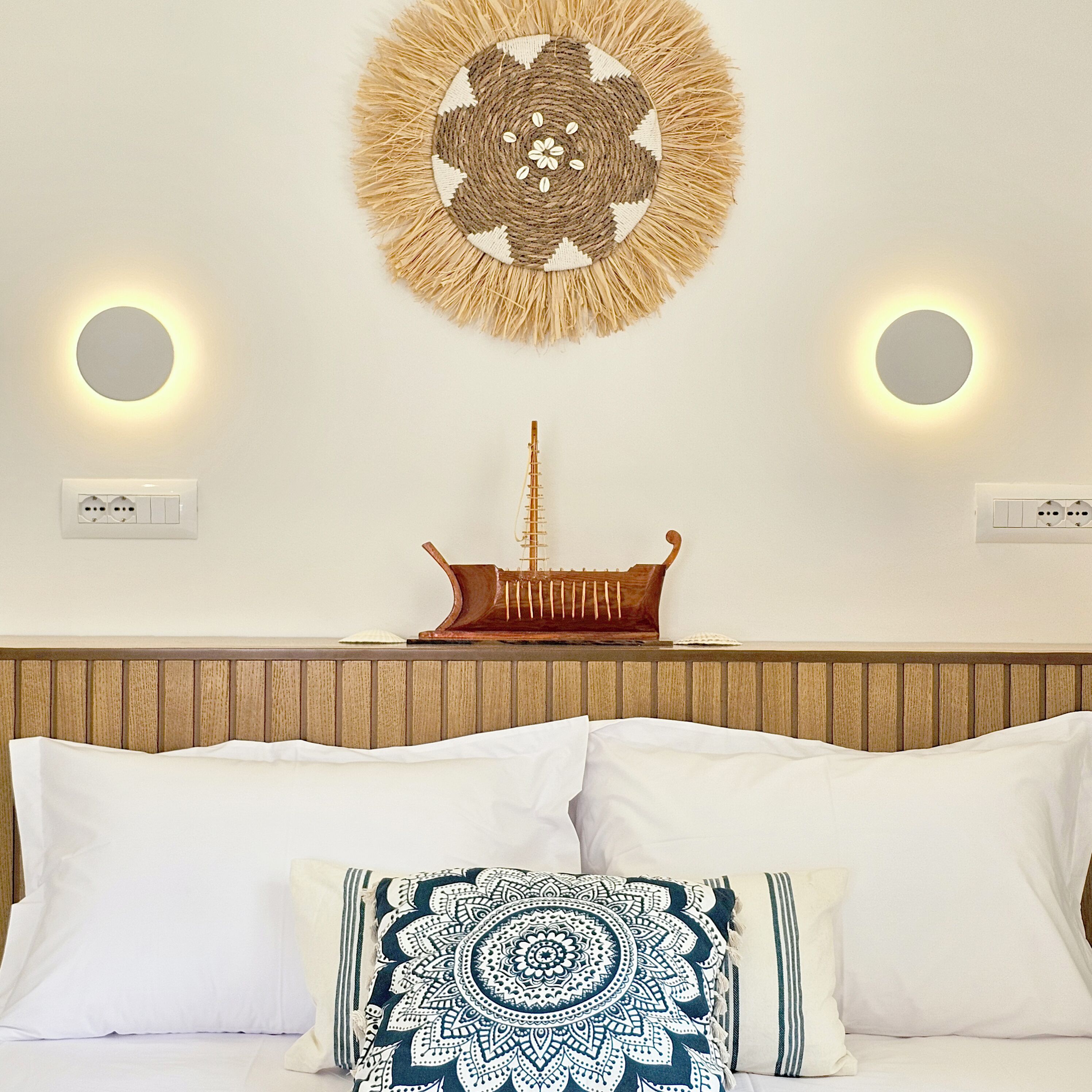 Een slaapkamer met een houten hoofdbord, een decoratieve wandhanger, twee wandlampen en een bed met wit beddengoed en een kussen met een patroon.
