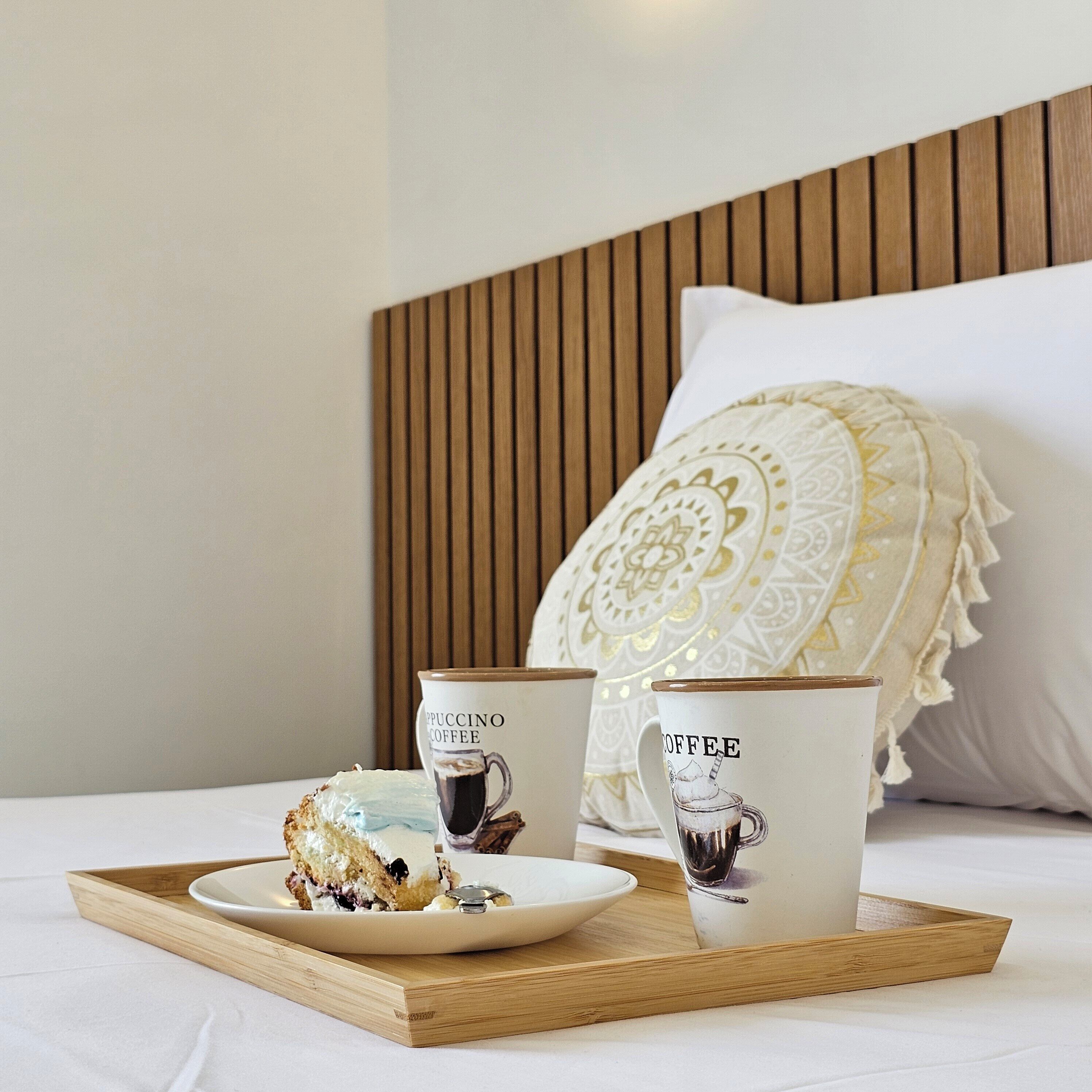 Un lit avec une tête de lit en bois, un oreiller blanc et un plateau sur lequel se trouvent une part de gâteau et deux tasses à café.
