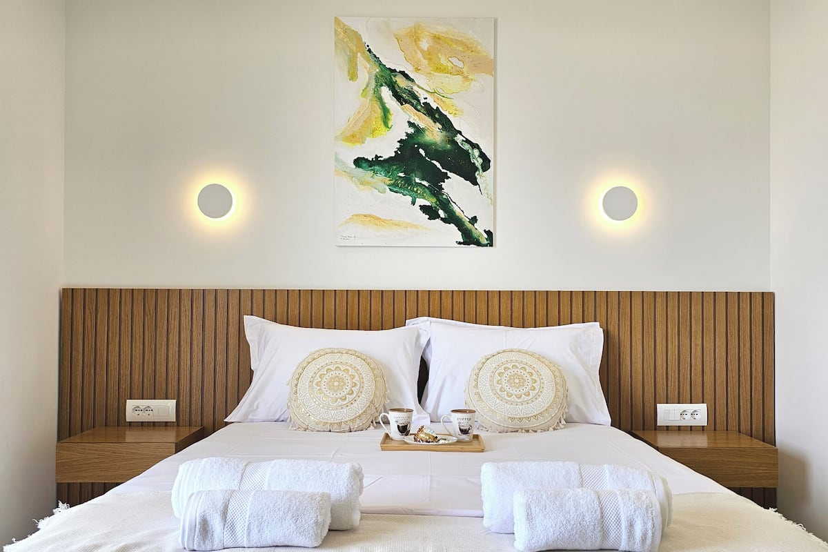 Una cama bien hecha con sábanas blancas, dos almohadones decorativos y una bandeja con una taza de café y un plato de comida.