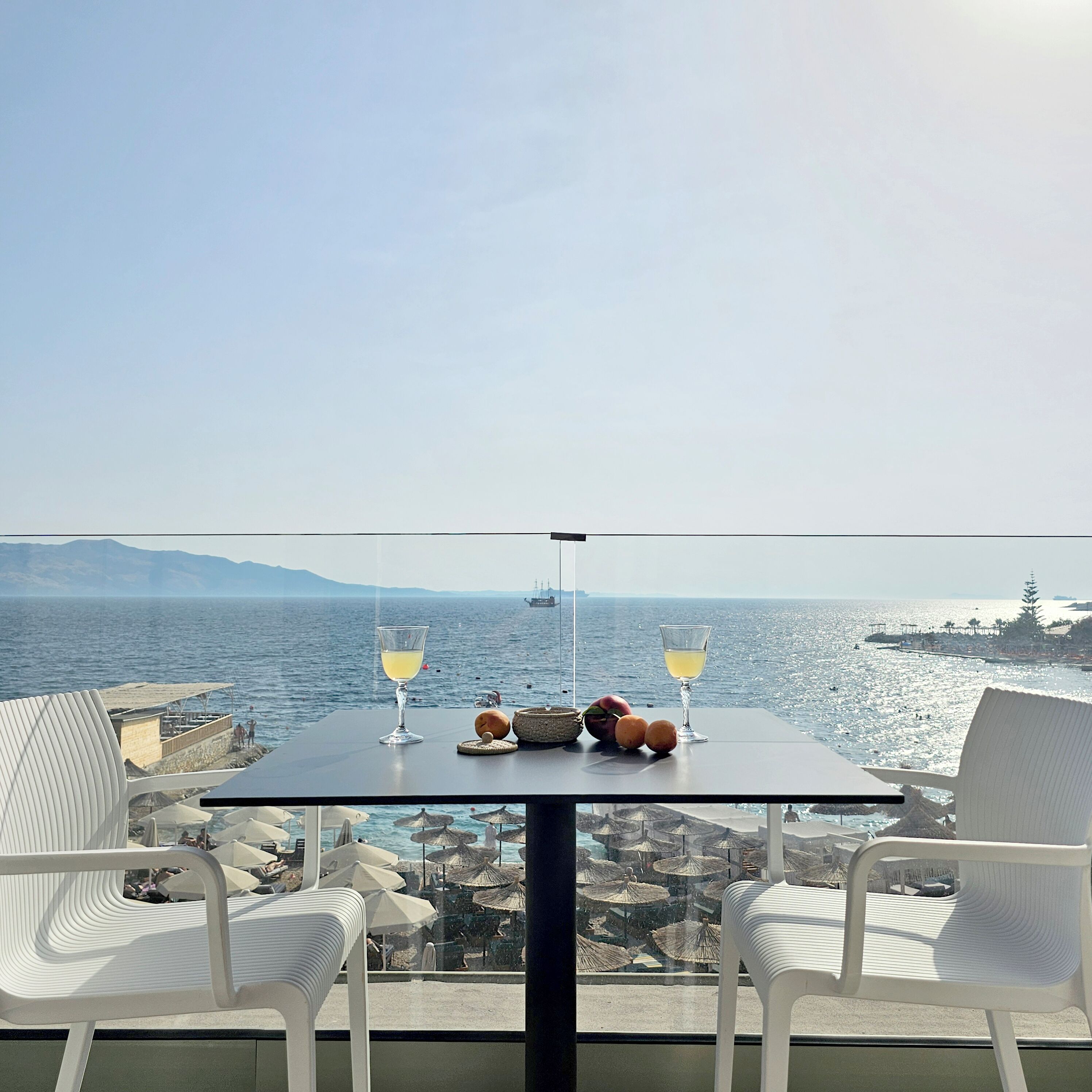 Une table avec deux verres d’une boisson jaune, un panier et quelques fruits, sur un balcon donnant sur une plage avec des parasols et des gens.