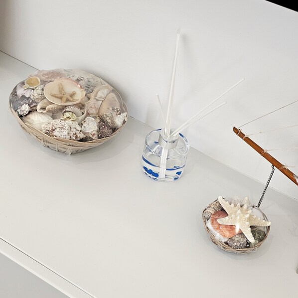 A table with a decorative bowl containing seashells and starfish, a clear glass bottle with reed diffusers, and a small basket with a starfish and seashells. - Ares Beachfront Luxury Rooms (Sarande)