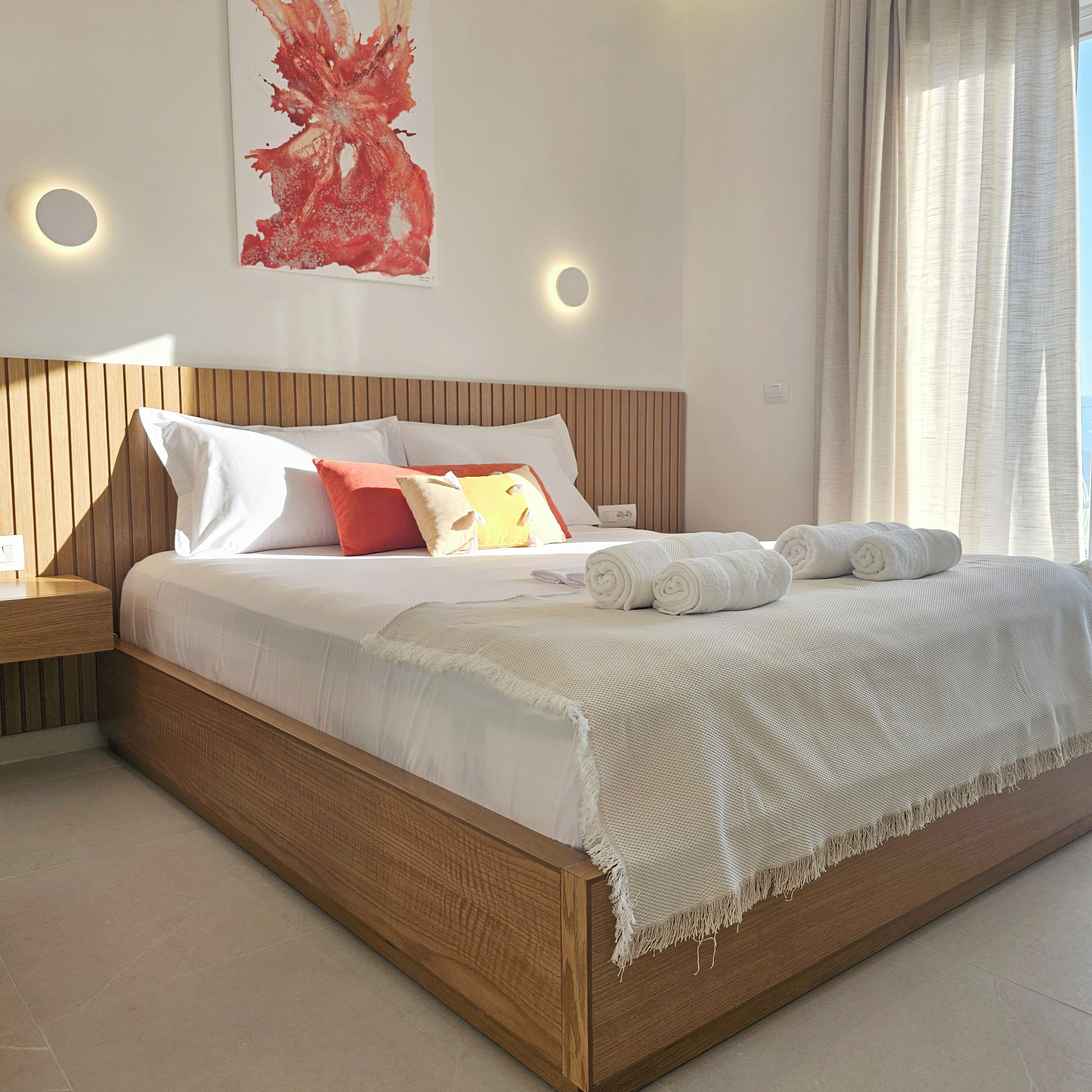 A modern bedroom with a wooden headboard, white bedding, and a large abstract painting on the wall.