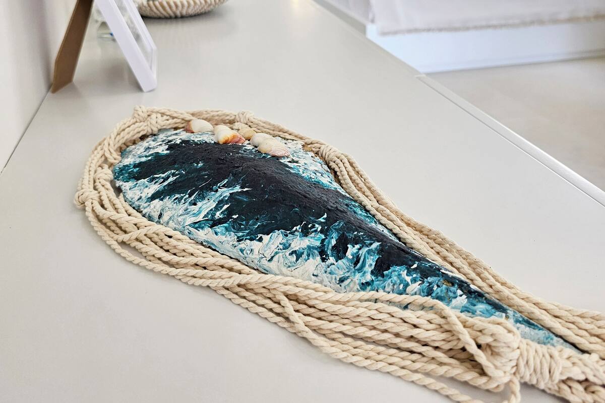 Una habitación con una cama, una mesita de noche con un colgador de macramé y un cuadro en la pared.