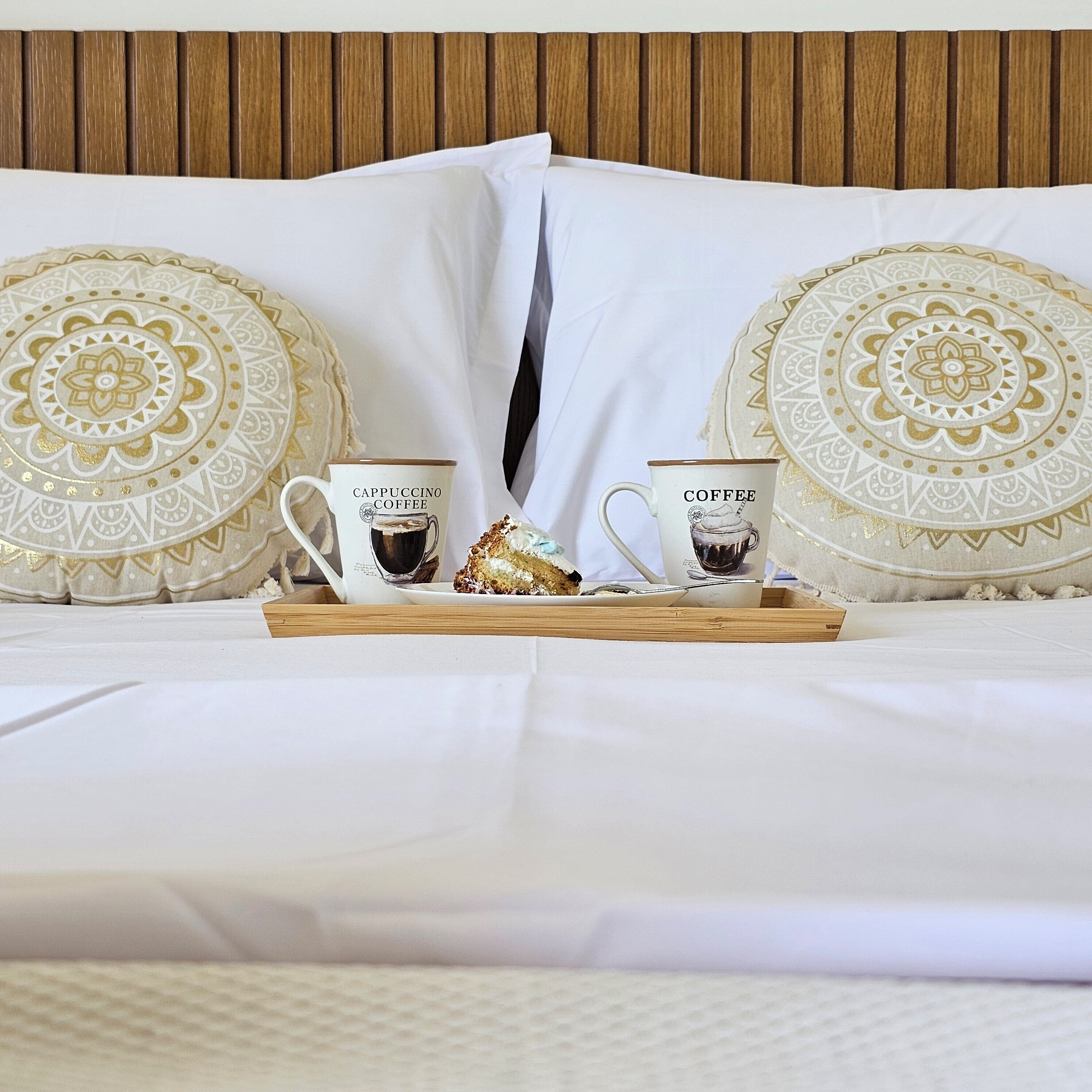 A neatly made bed with two decorative pillows, a tray with two coffee mugs and a pastry, and a wooden headboard.