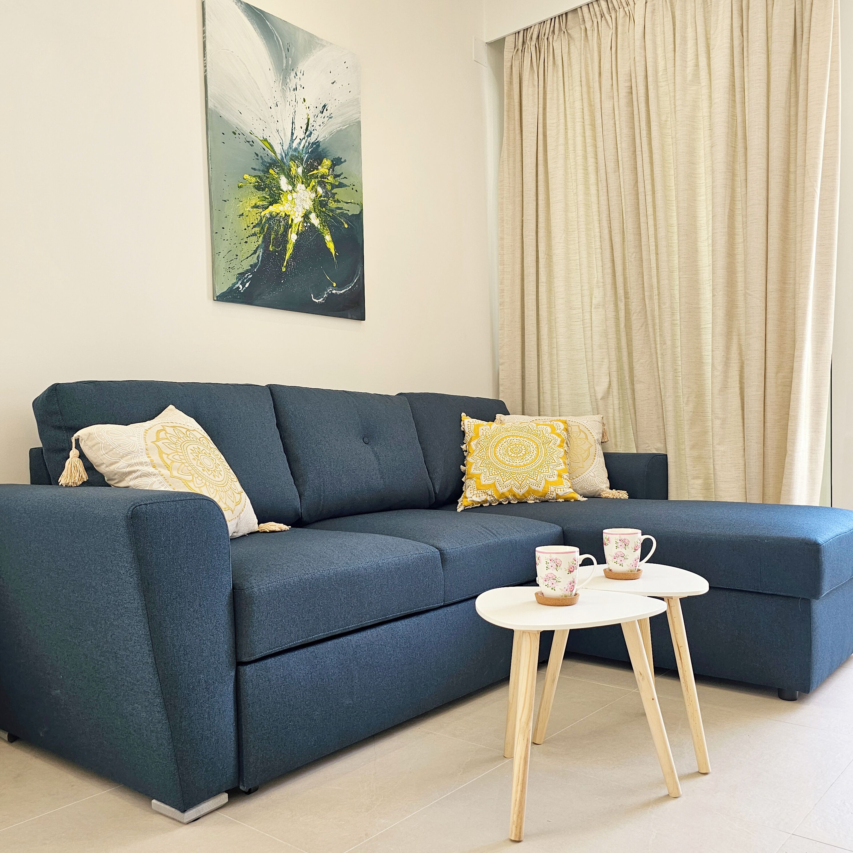 Un salon moderne avec un canapé bleu foncé, deux coussins décoratifs, une petite table blanche sur laquelle reposent deux tasses à motifs floraux, et un grand tableau abstrait accroché au mur.