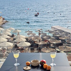A table with two glasses of a yellow beverage, a basket with fruits, and a view of the sea with beach umbrellas and lounge chairs.