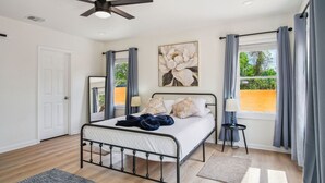 A bedroom with a bed, a ceiling fan, a window with curtains, a painting, and a door. - Stylish Spacious Comfort 4BR 3BA Downtown Gem (San Antonio)