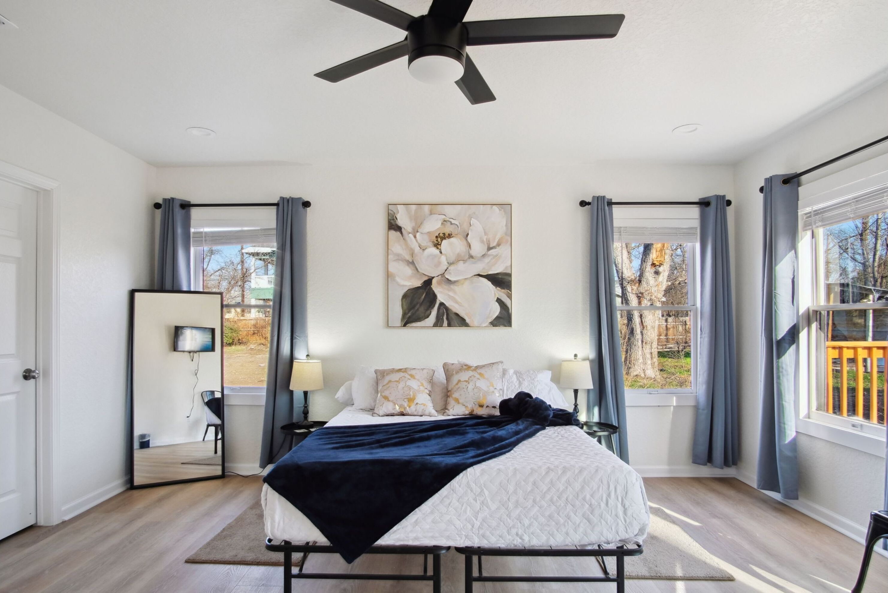 Une chambre à coucher avec un lit, un ventilateur de plafond, une grande fenêtre avec des rideaux et un téléviseur fixé au mur.