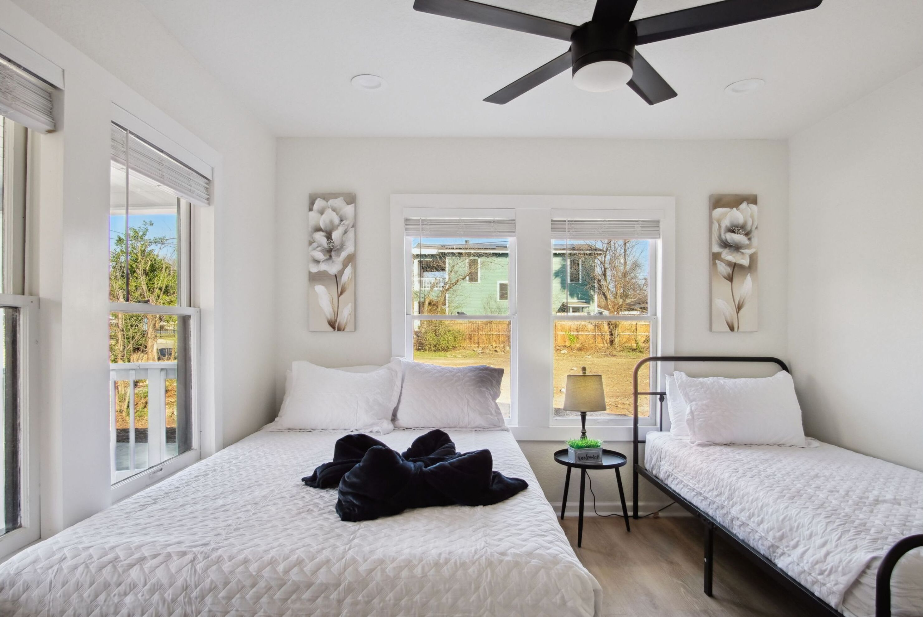 A bedroom with two beds, a ceiling fan, and large windows with views of greenery and buildings.