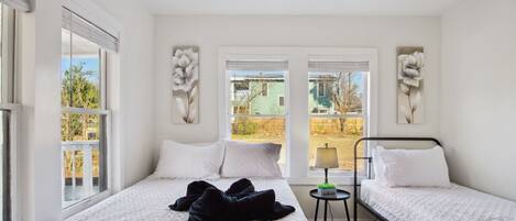 A bedroom with two beds, a ceiling fan, and large windows with views of greenery and buildings.