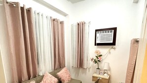 Een moderne woonkamer met een bank, zachte roze kussens, een rond vloerkleed, een bureau met stoel, een bijzettafeltje met lamp en een wandairconditioner.