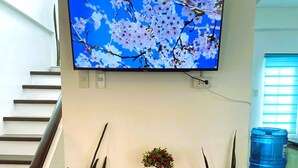 Een woonkamer met een televisie aan de muur, een klein tafeltje met een vaas en een rode map, en twee potplanten.