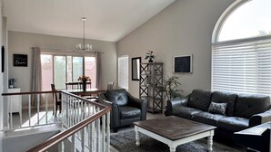 Un vaste salon doté d’un haut plafond, un coin repas avec une table et des chaises, un canapé en cuir, une table basse et un escalier avec des rampes blanches.