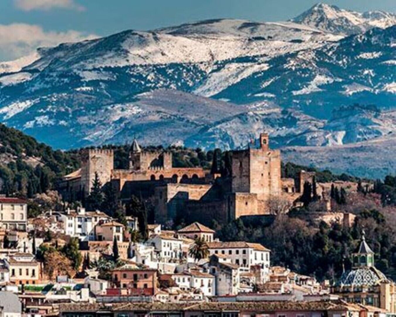 A historic town with a prominent castle on a hill, surrounded by buildings and a mountainous backdrop.