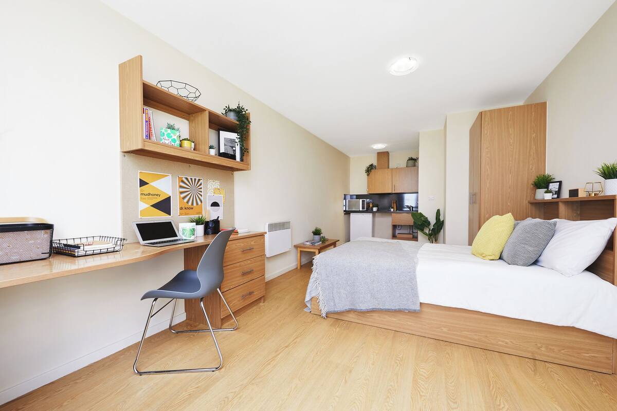 Un dormitorio moderno con un escritorio de madera, una cama con almohadas y una kitchenette al fondo.