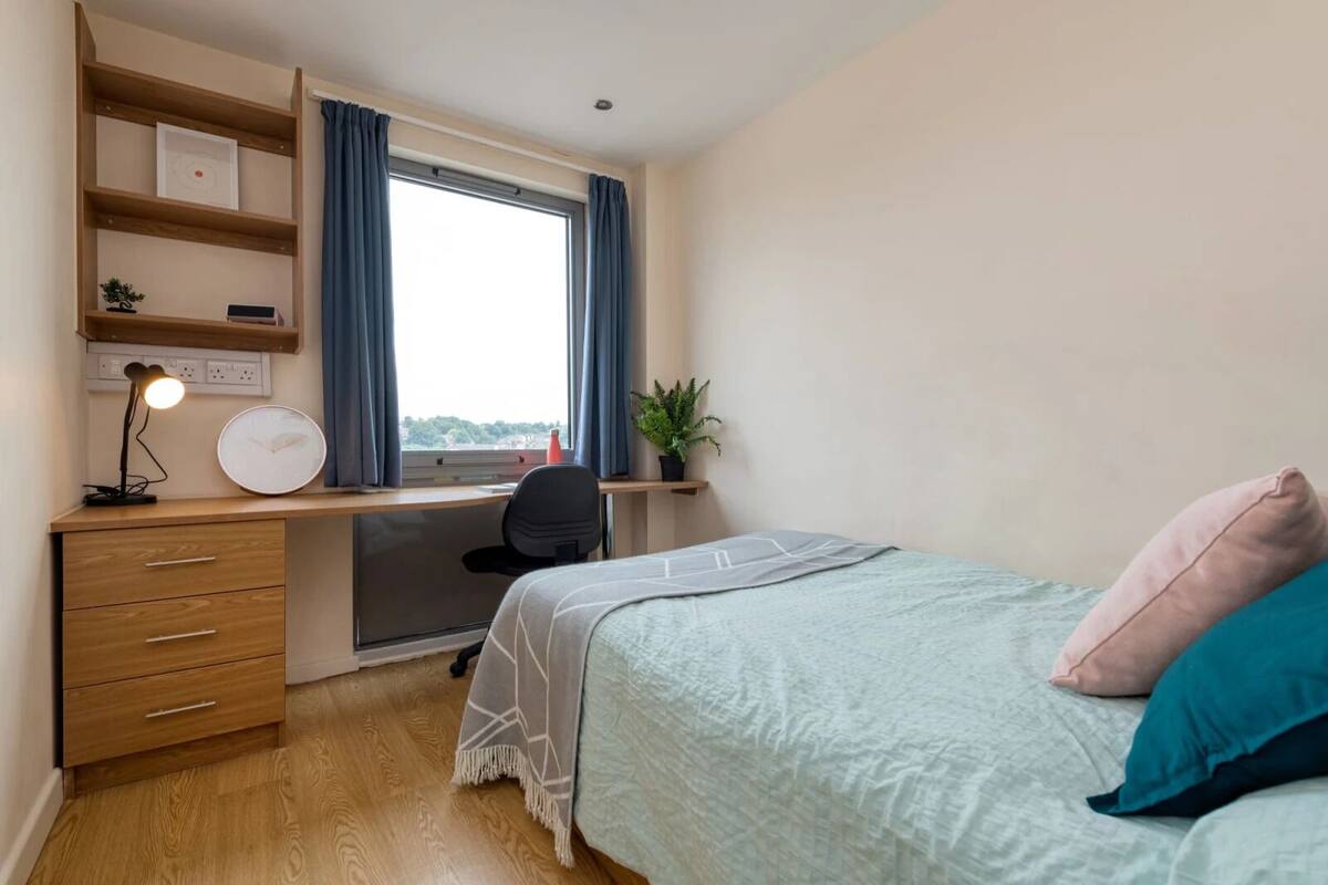 Un dormitorio con cama, escritorio, silla, lámpara y ventana con cortinas.