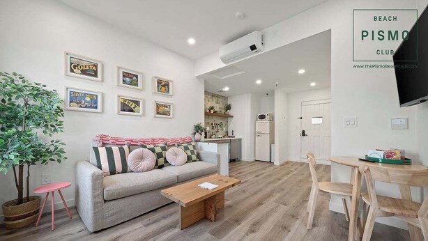 Una sala de estar moderna con sofá, mesa de centro y comedor. Hay cuadros enmarcados en la pared, una planta en maceta y un televisor montado.