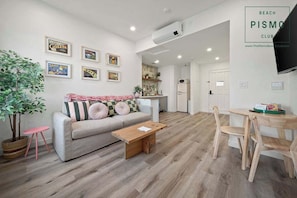 Una sala de estar moderna con sofá, mesa de centro y comedor. Hay cuadros enmarcados en la pared, una planta en maceta y un televisor montado.