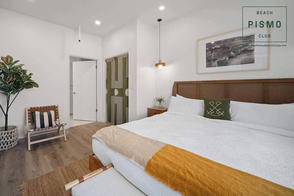 Une chambre avec un grand lit, une table de chevet, une lampe suspendue, une chaise, une plante et une porte ornée d’un motif géométrique.