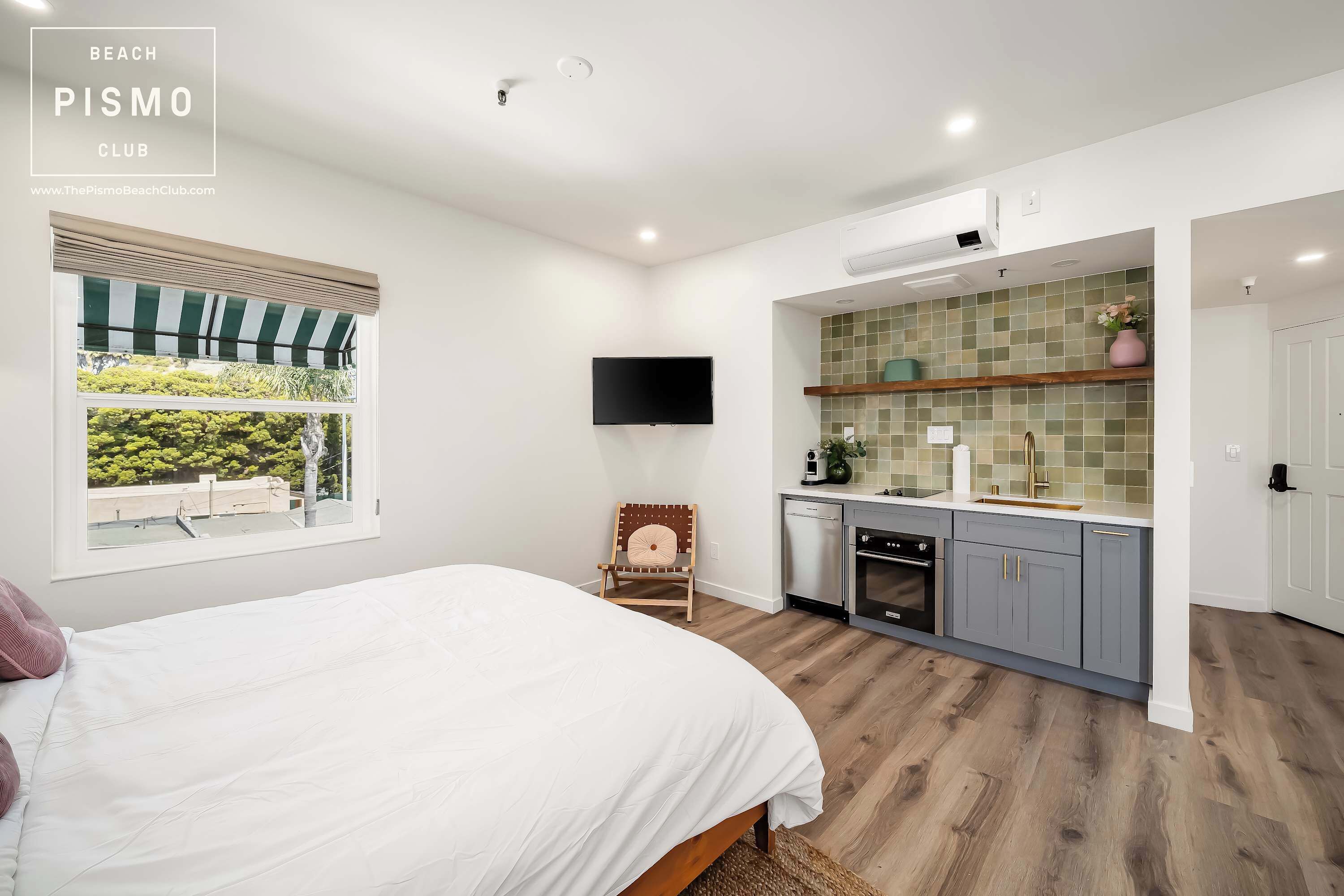 A bedroom with a bed, a window with a view, a chair, a TV, a kitchenette with cabinets, and a tiled backsplash.