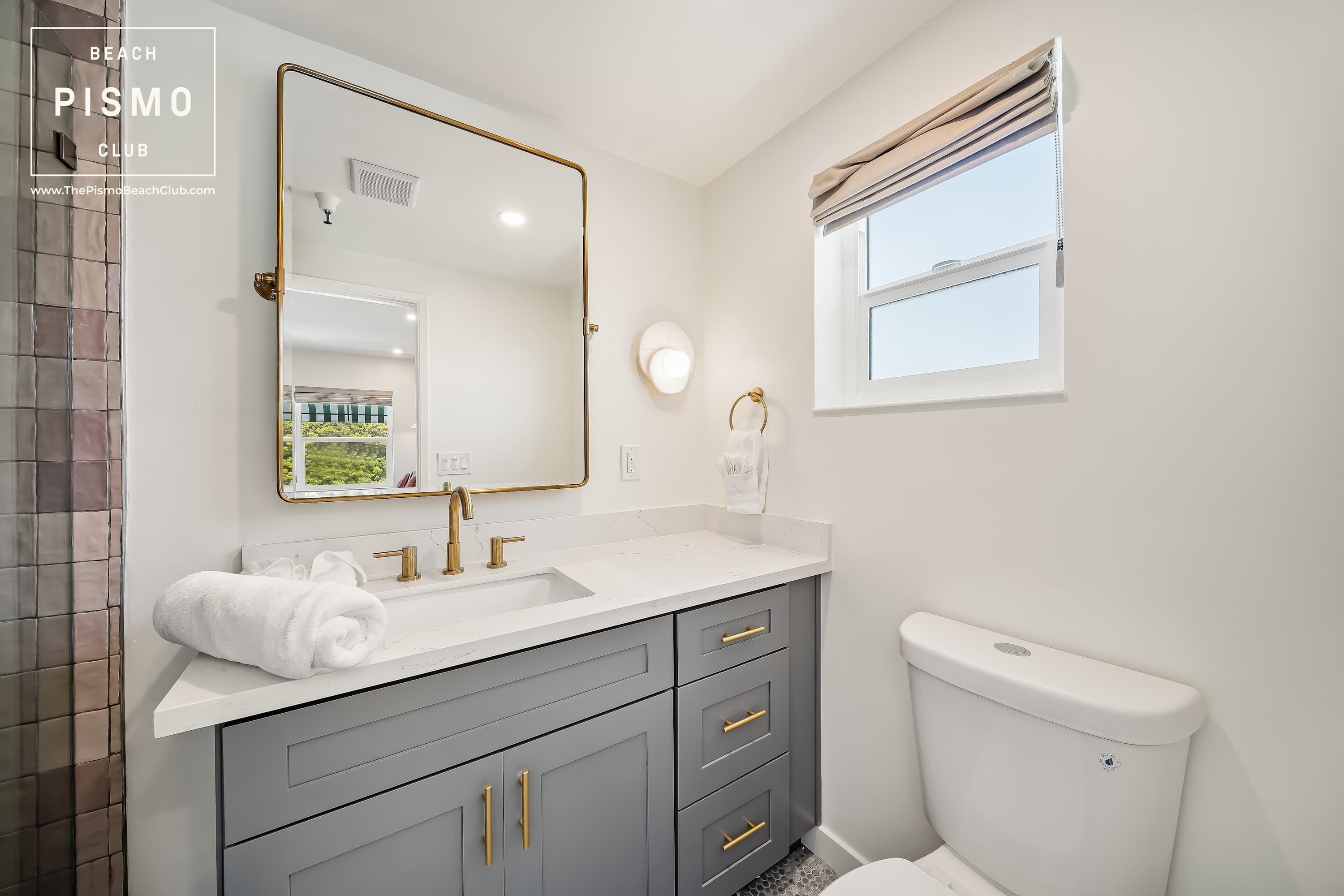 A bathroom with a white sink, grey cabinets, a gold-framed mirror, and a window with beige curtains.