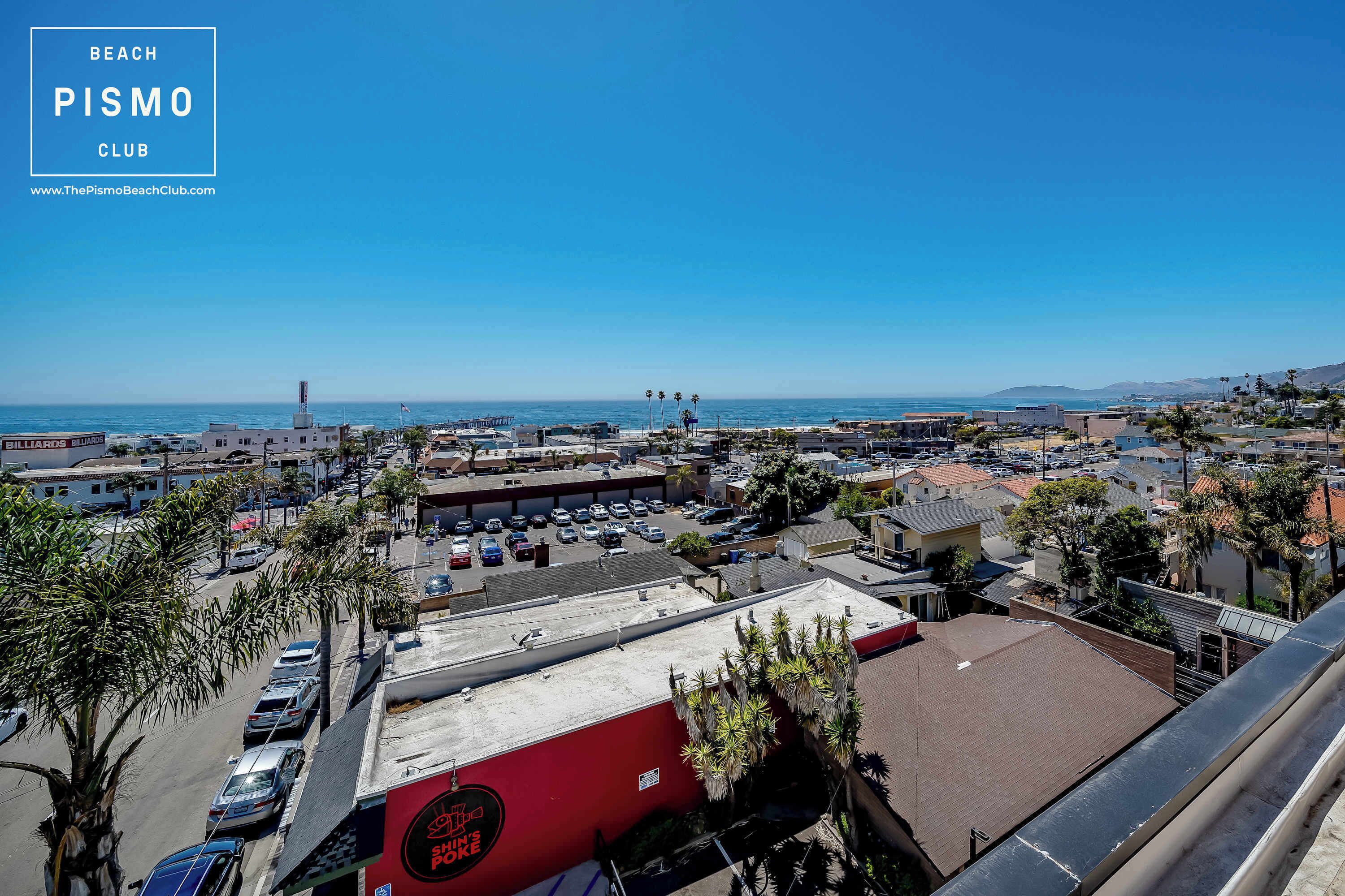 A coastal cityscape with a clear view of the beach, a parking lot, and a building with a red awning.