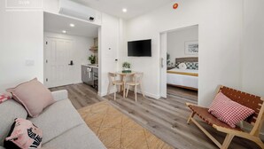 Un salon moderne avec un canapé, un banc en bois, un coin repas avec une table et des chaises, un téléviseur fixé au mur et une cuisine en arrière-plan.