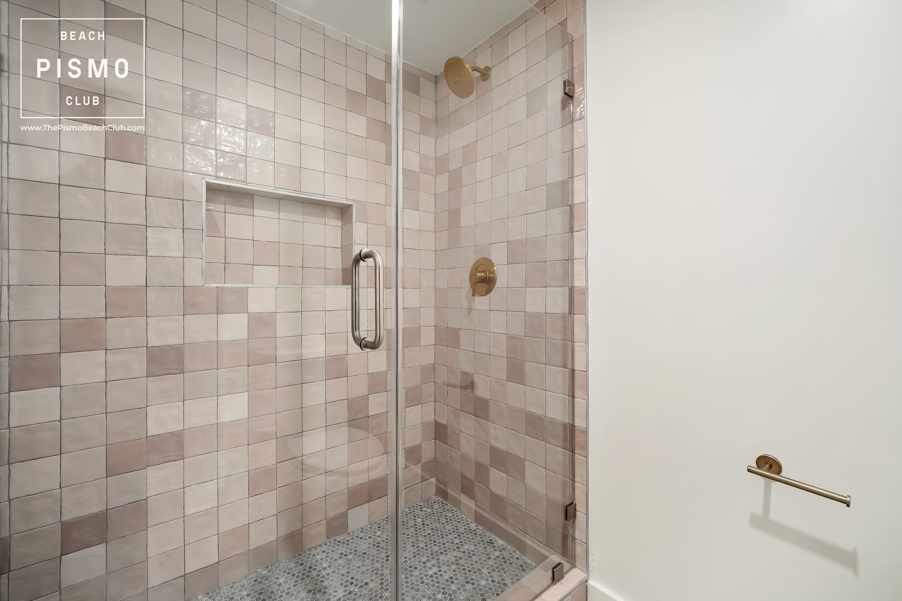 Eine Dusche mit Glastür, beige- und rosafarbenen Fliesen und einem goldfarbenen Duschkopf.