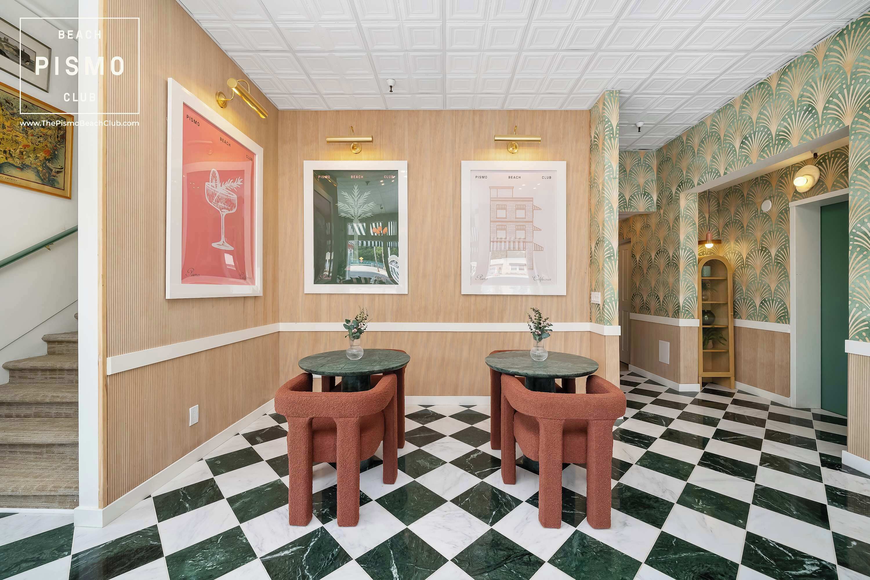 Un couloir doté d’un sol à carreaux, deux tables rondes avec des chaises, des affiches encadrées accrochées au mur et un escalier sur le côté gauche.