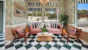 Une pièce avec un sol à carreaux, un canapé rose, deux fauteuils en bois, une table basse en bois et des auvents à rayures au-dessus des fenêtres.