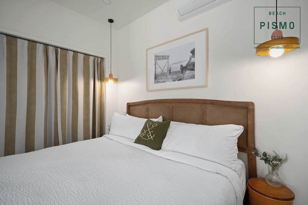 Een keurig opgemaakt bed met wit beddengoed en één groen kussen, een ingelijst schilderij aan de muur, en een klein houten nachtkastje met een vaas.