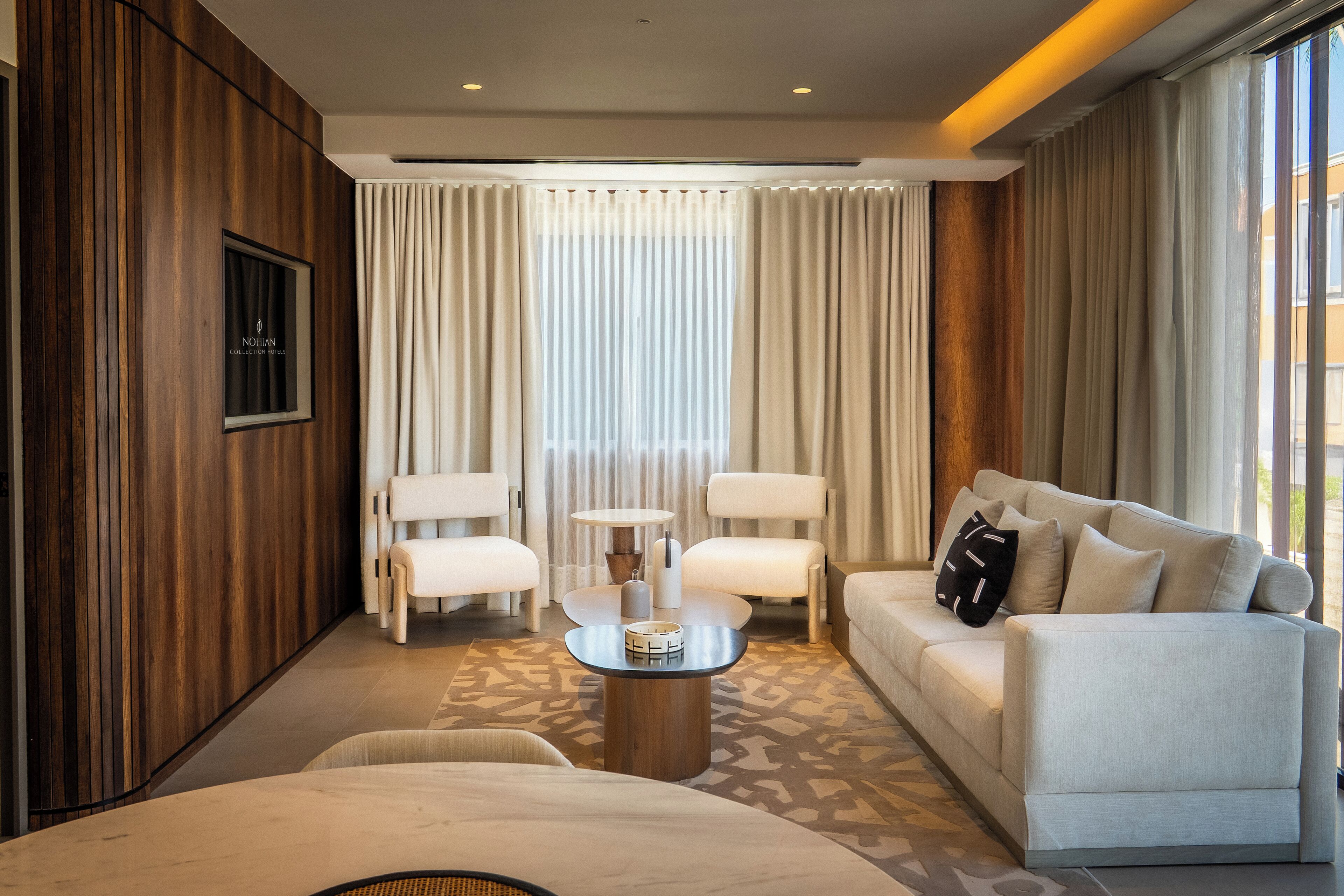 Una habitación de hotel moderna con sofá, dos sillones, una mesa de centro y un ventanal con cortinas.