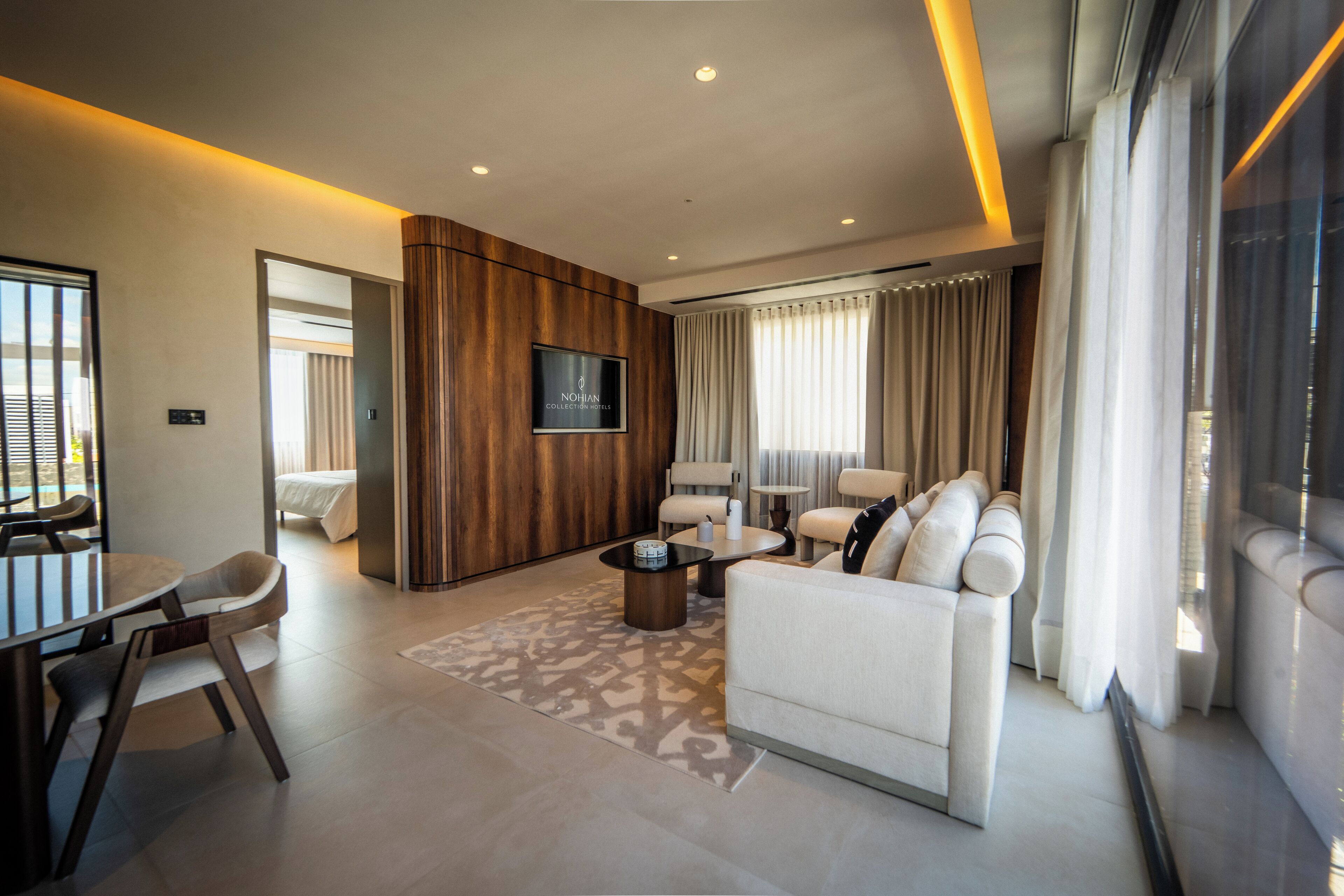 A modern hotel room with a sofa, a dining area, a TV, and a large window with curtains.