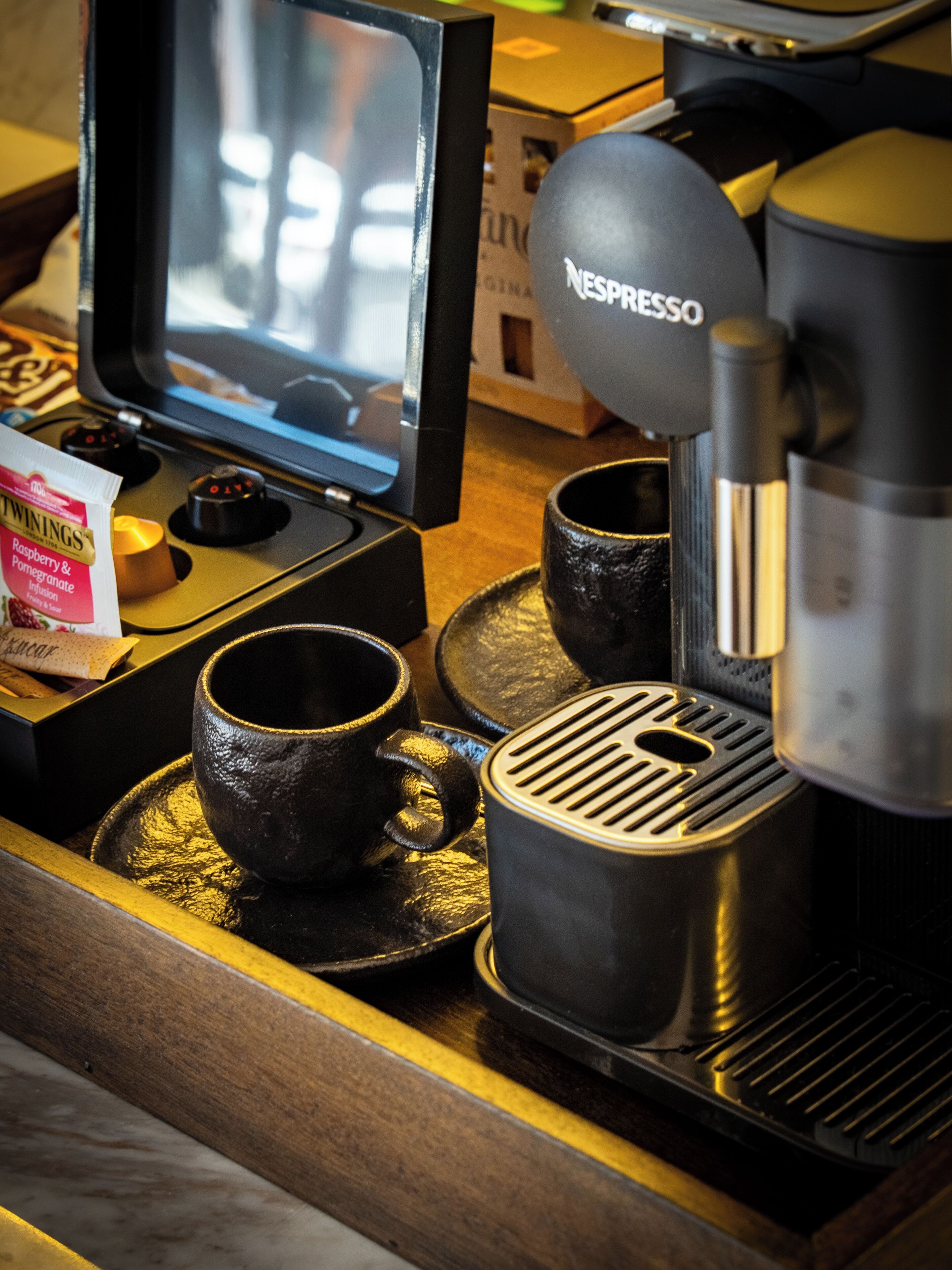 Una máquina de café con dispensador para cápsulas Nespresso, dos tazas negras, una caja de café Twinings y una pequeña pantalla de televisión.