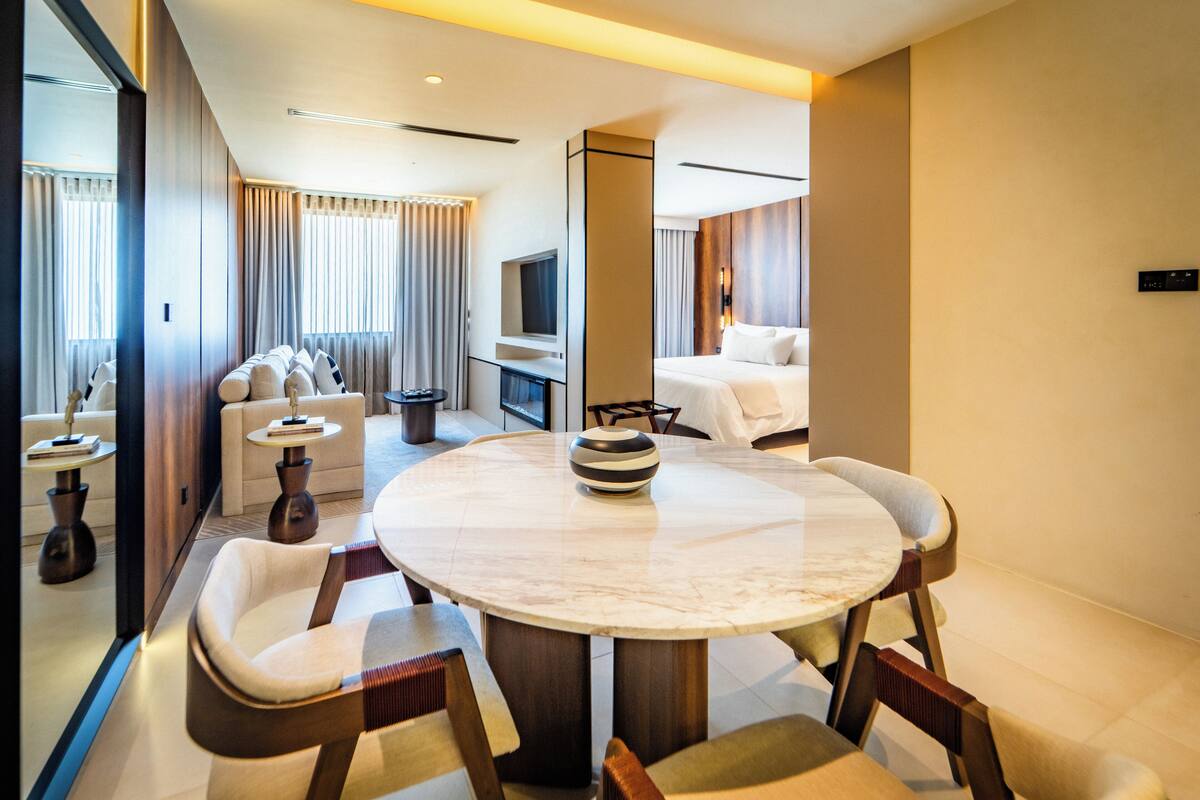 A modern hotel room with a round marble dining table, two sofas, a TV, and a minibar.