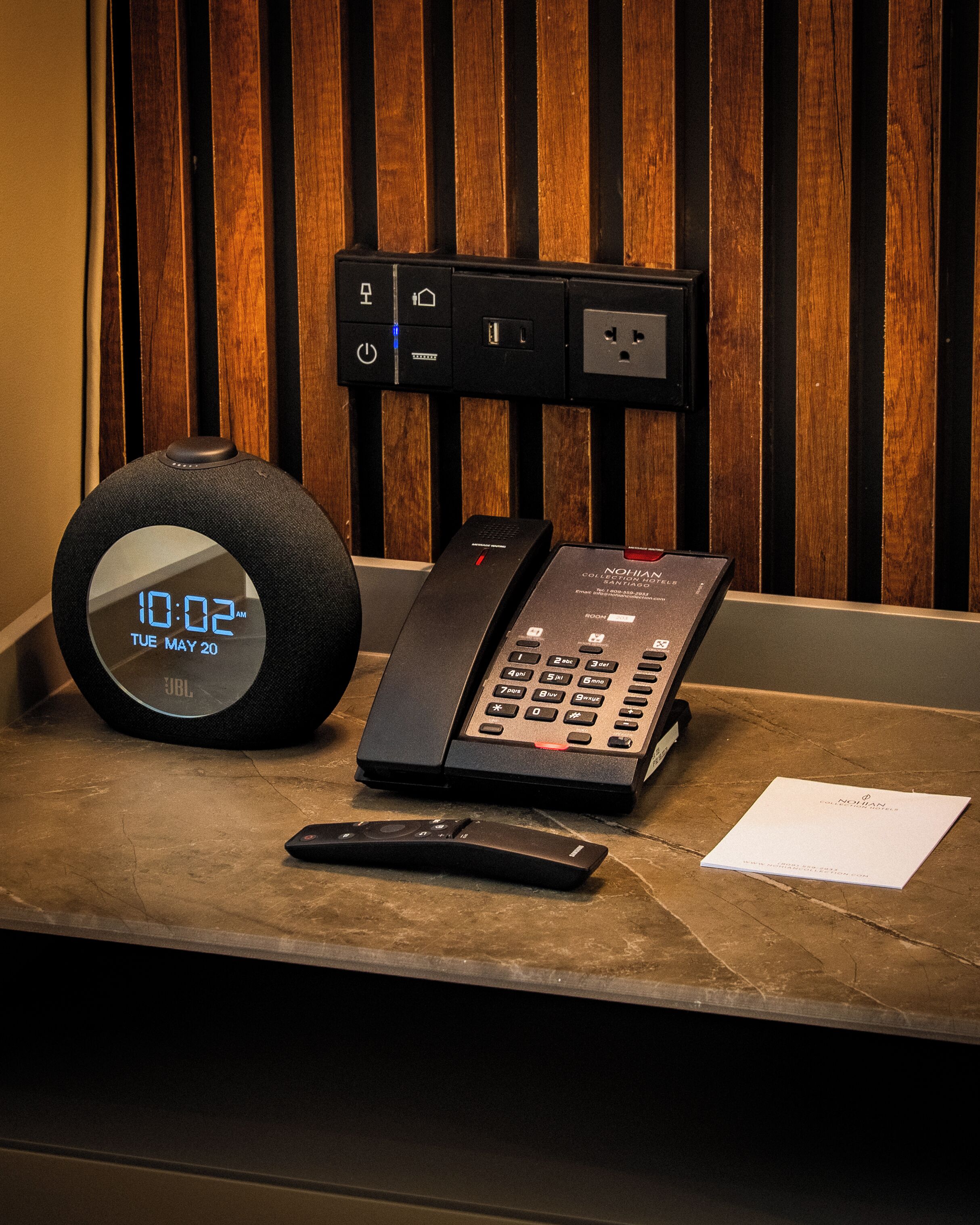 A digital clock displaying 10:02 AM, a telephone, and a remote control on a wooden surface.