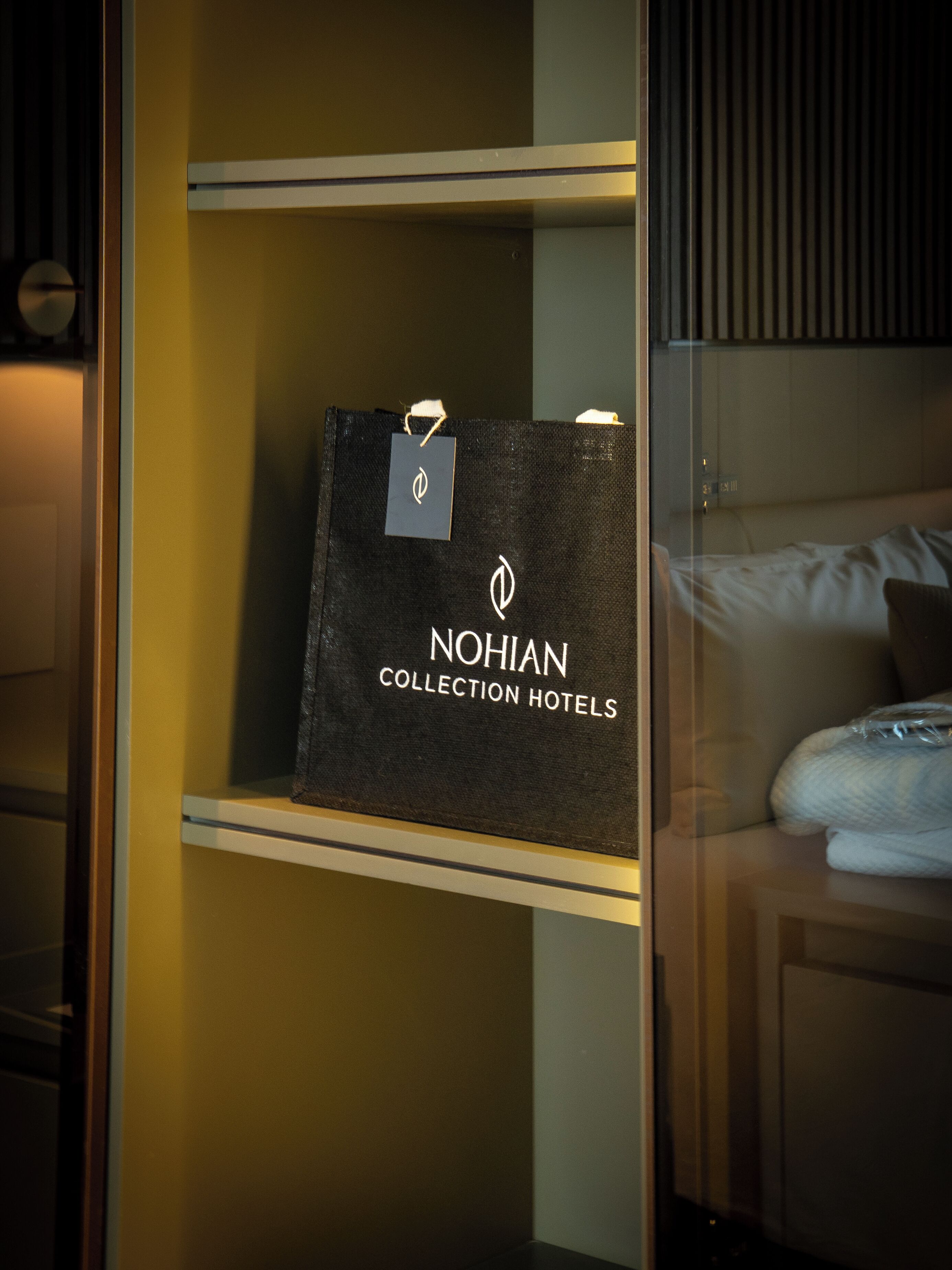 Eine schwarze Tasche der NOHIAN COLLECTION HOTELS wird in einer Vitrine präsentiert, zusammen mit einem Anhänger, in einem Zimmer mit Bett und hölzernem Kopfteil.