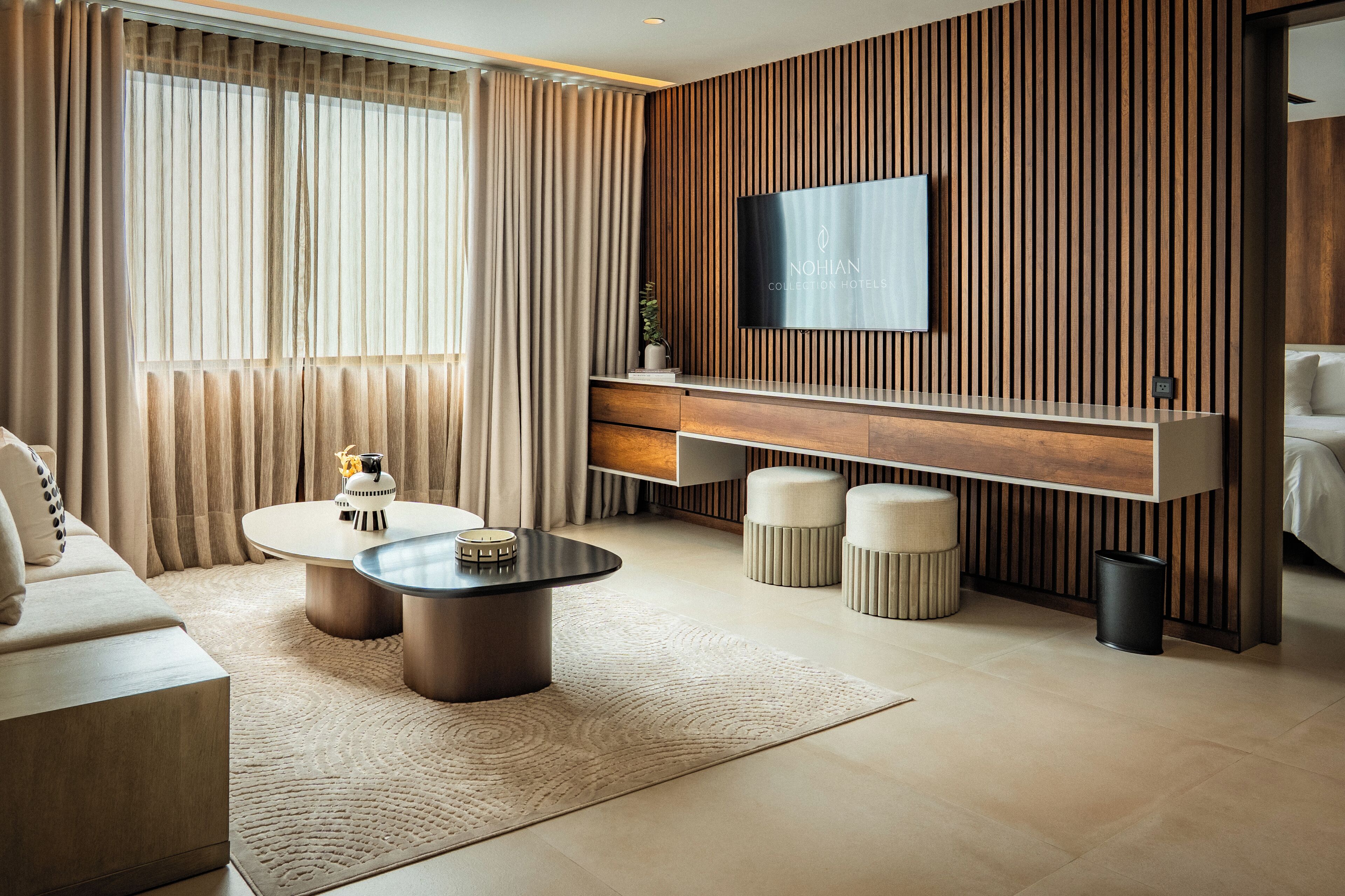 A modern hotel room with a large flat-screen TV mounted on a wooden panel wall, a round coffee table, a sofa, and a small side table with a decorative item.