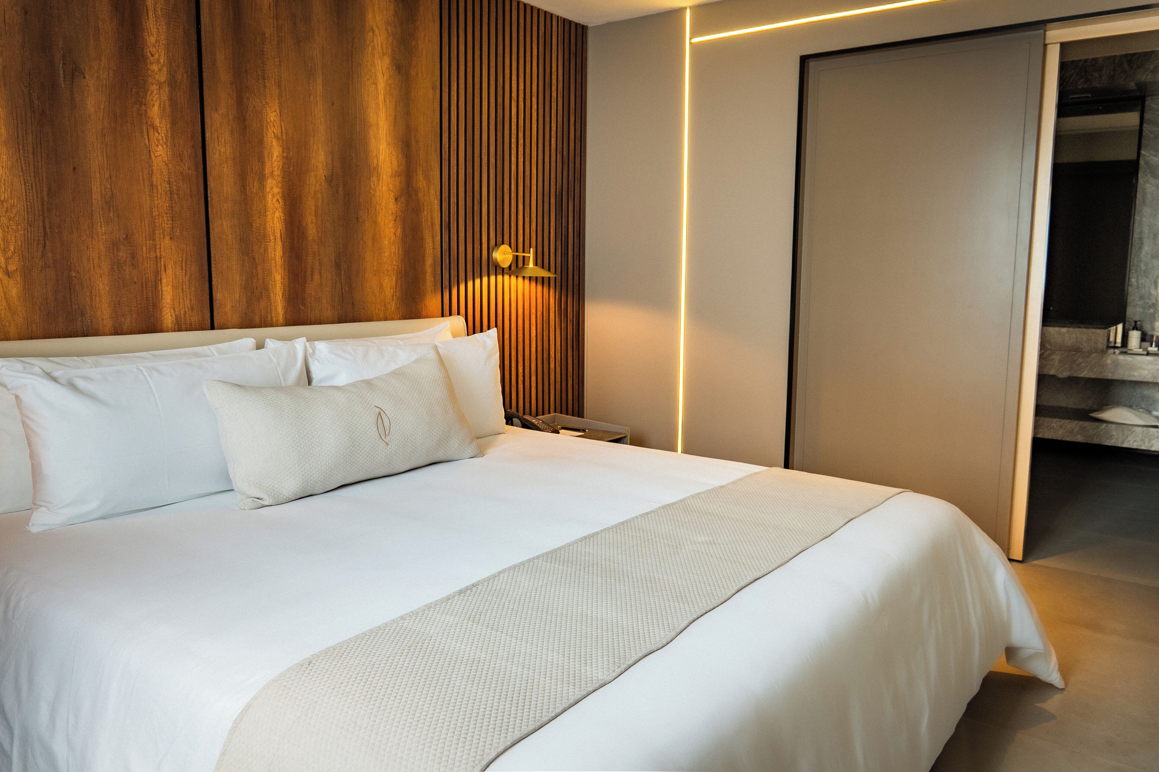 Quarto de hotel moderno com uma cama grande, painéis de madeira na parede, uma mesa de cabeceira e um banheiro visível através de uma porta aberta.