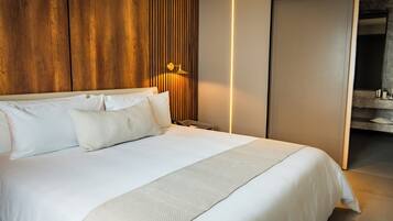 Quarto de hotel moderno com uma cama grande, painéis de madeira na parede, uma mesa de cabeceira e um banheiro visível através de uma porta aberta.