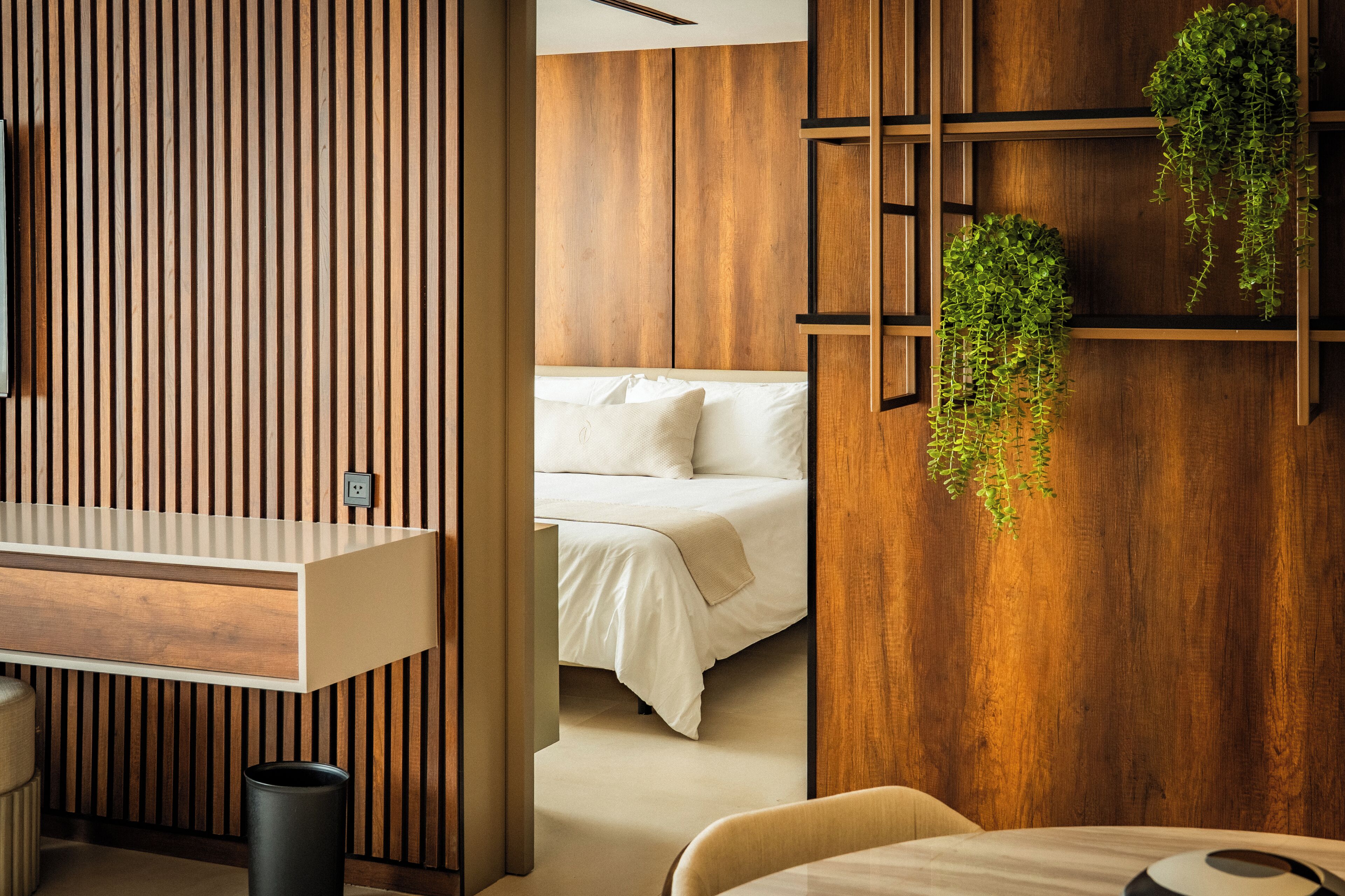 A modern hotel room with a wooden slat wall, a bed with white bedding, a bedside table, and a hanging plant.