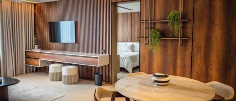 A modern hotel room with a wooden accent wall, a flat-screen TV, a bed, a dining area with a round table and chairs, and a small plant on the wall.