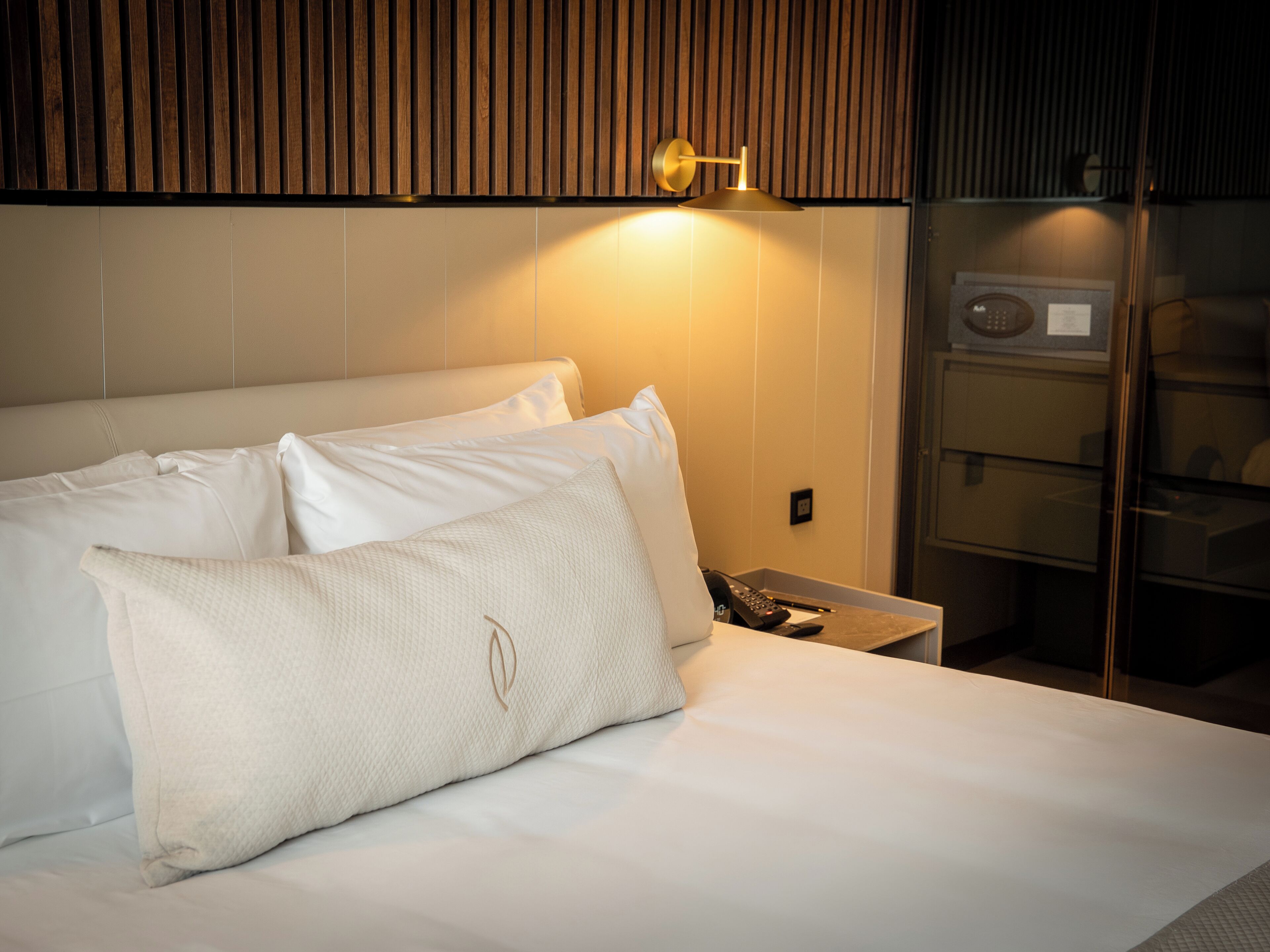 A neatly made bed with white linens and two pillows, a bedside table with a phone and a remote control, and a wall-mounted light fixture.