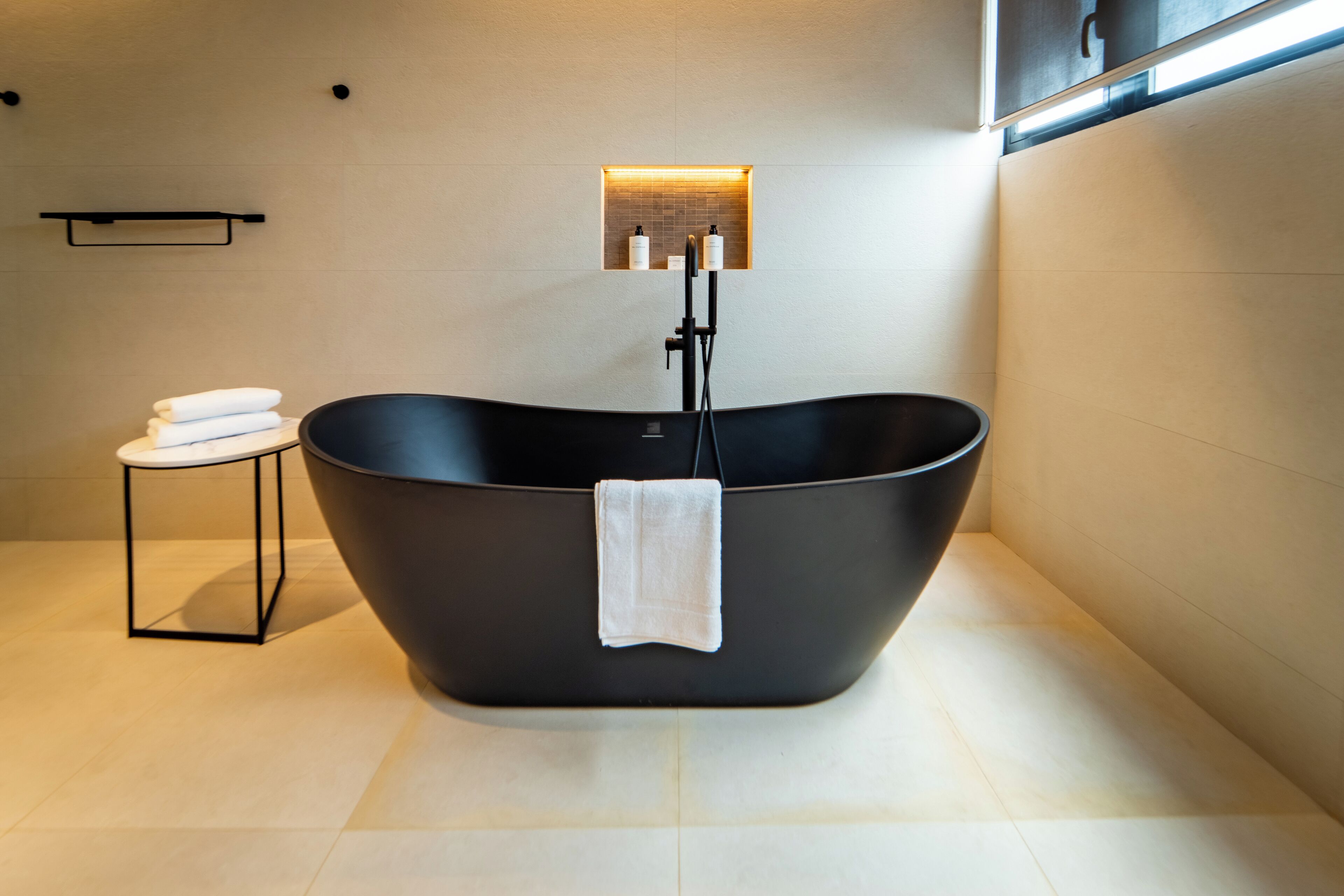 A modern bathroom with a freestanding bathtub, a small table with towels, and a wall-mounted shelf with toiletries.