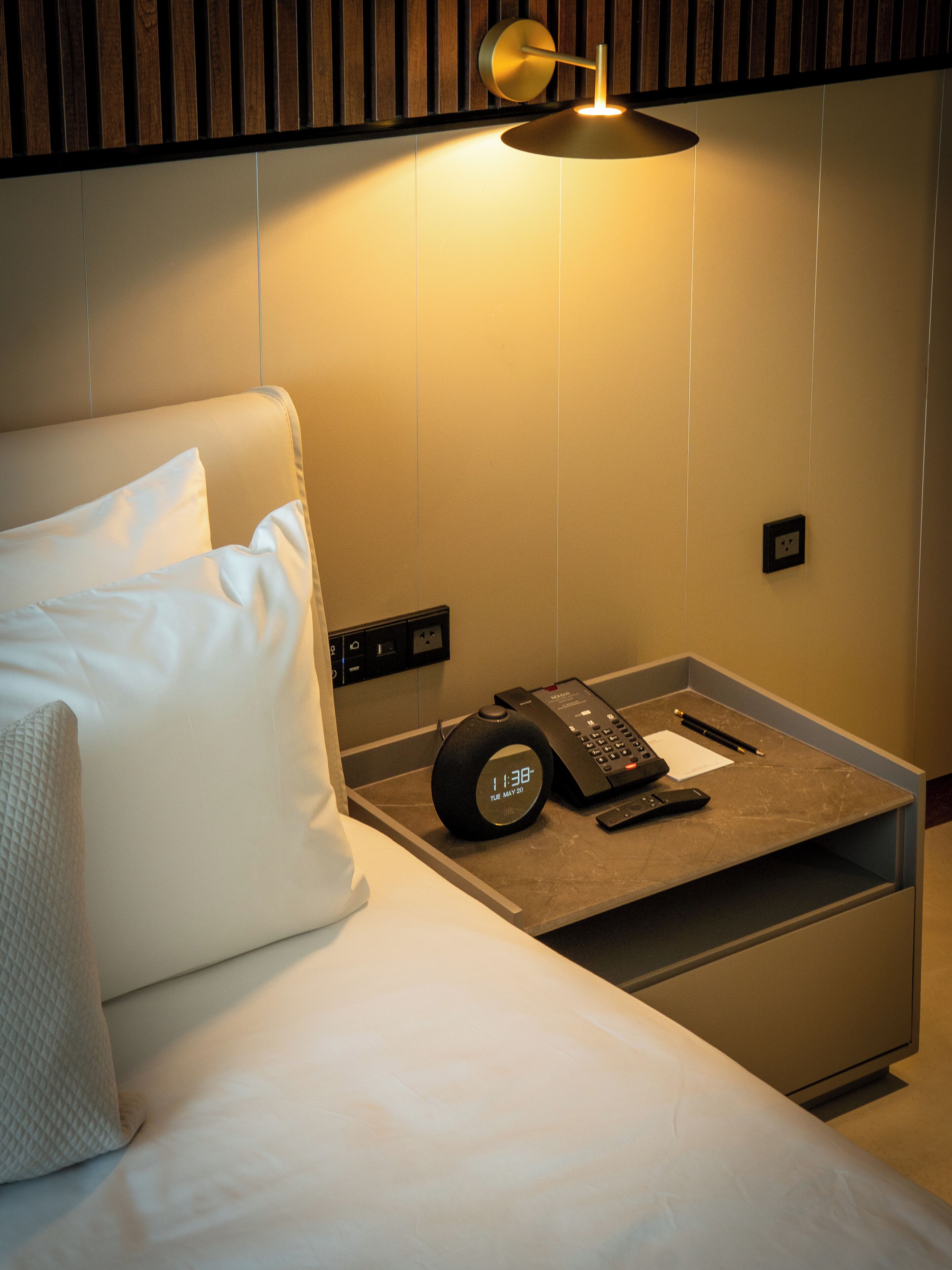 A hotel room with a bed, a bedside table with a phone and a remote control, and a wall-mounted light fixture.